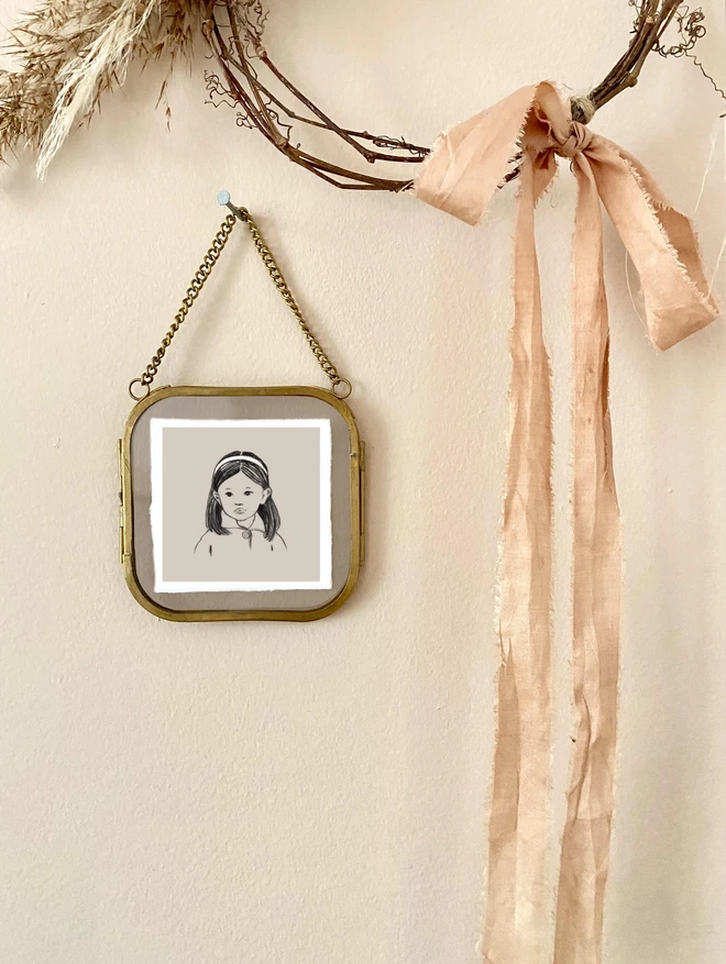 a photograph of a miniature drawing of an asian Japanese girl with a white head band framed in a small glass fame hug on a wall next to hand made wreath