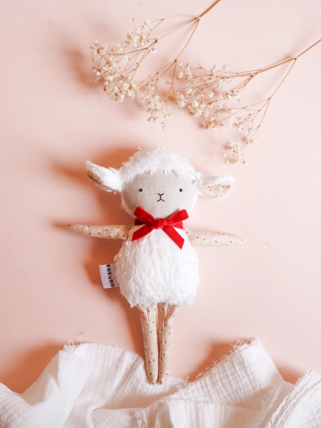 Handmade lamb doll with a fluffy white body, stitched face, beige floral fabric arms and legs, and a small red velvet bow tied around its neck.