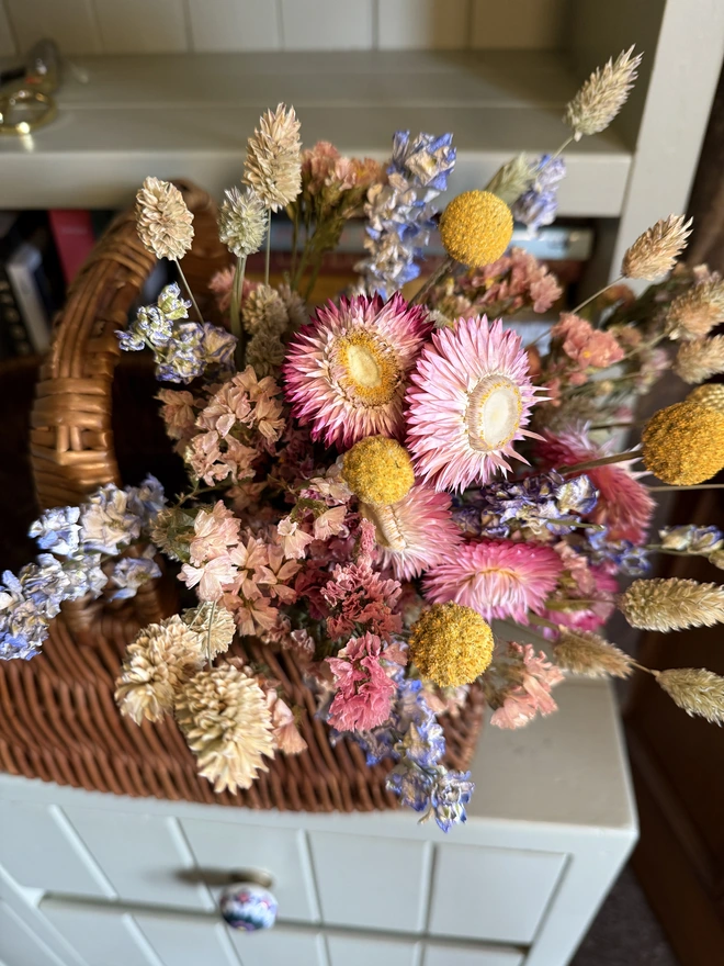 a bouquet of dried flowers in bright colours displayed in a wicker basket against a green painted dresser