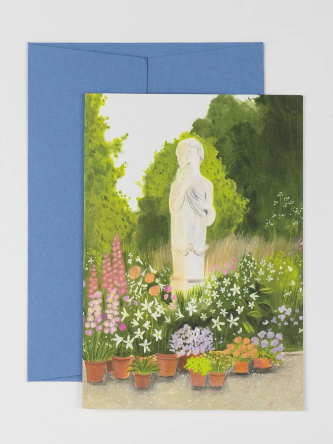 A white statue of a woman standing amongst colourful flower pots. The sun filters through the trees in the background to give a glowy golden light