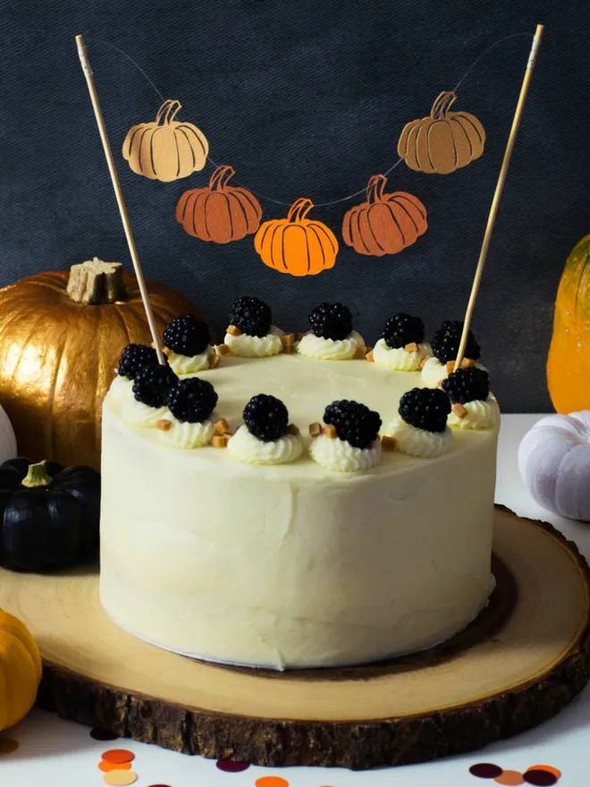 Cake bunting with 5 mini paper pumkins suspended between two bamboo skewers. Cake bunting is inserted into a cake decorated with blackerries on a party table decorated with pumpkins.