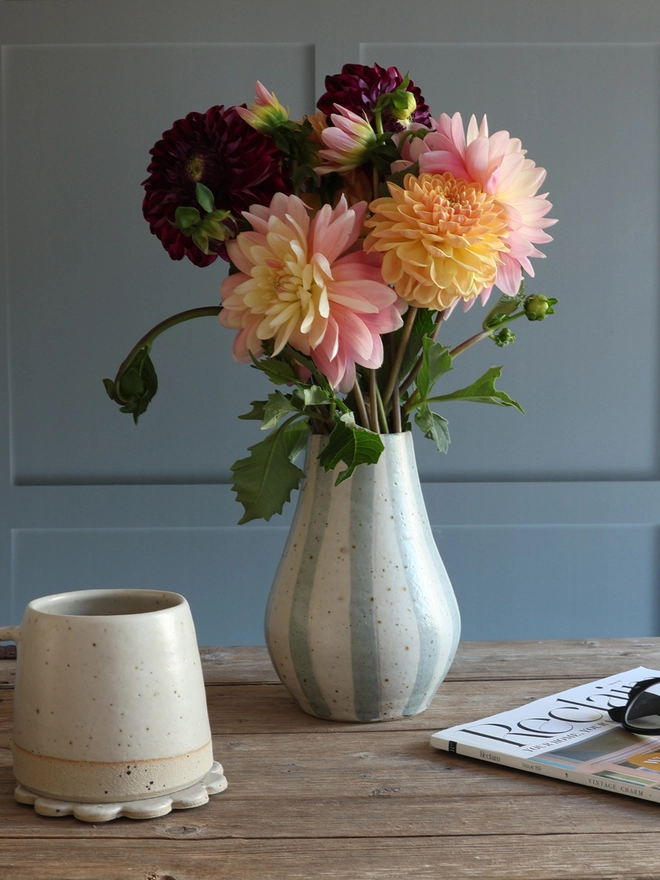Large Handmade Ceramic Stoneware Vase in Blue and White Stripe holding a bouquet of pink, yellow and red flowers. The vase sits on a wooden surface next to a handmade ceramic white mug sitting on a ceramic coaster, and a magazine with a pair of glasses on top. 