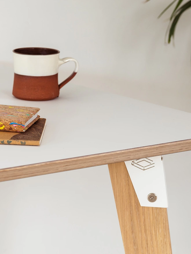 corner of a stylish and minimalist desk with white Fenix top, white coloured steel brackets and solid oak legs, with a mug and notebook on it.