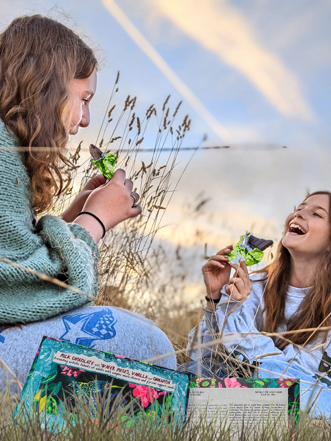 2 happy girls in a field enjoying charity milk chocolate wrapped in green foil & hummingbird packaging