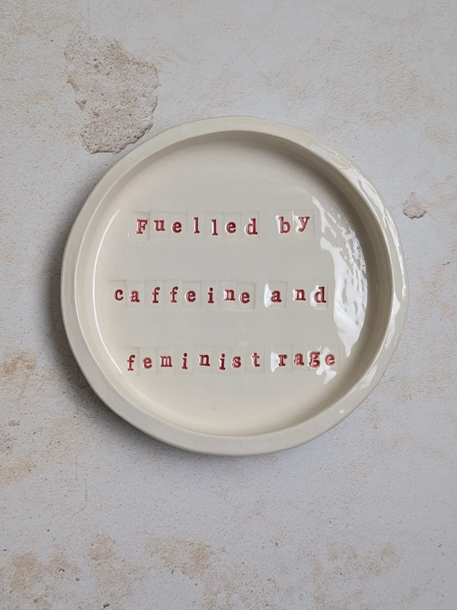 Fuelled by Caffeine and Feminist Rage a ceramic dish sitting on a grey surface. It is inscribed with a message in red writing. 