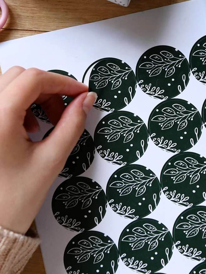 A hand is peeling a green botanical design sticker from a sheet of 35 matching stickers.