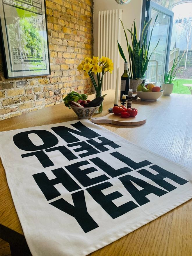 Hell Yeah Hell No black screen printed text on white tea towel laying flat on kitchen island with plant, daffodils in vase, fruit bowl, chopping board and bowl with vegetables in background