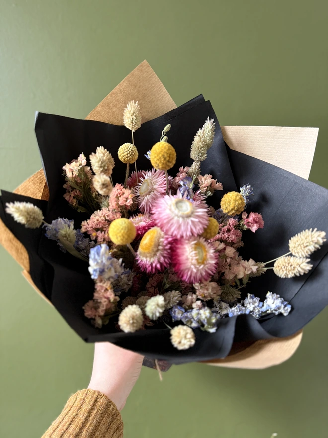 a bouquet of dried flowers in bright colours wrapped in black tissue and kraft paper held by a person in a mustard coloured jumper with their left arm against a green wall