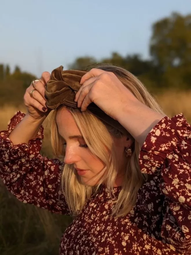 A caper green velvet fabric headband on a blonde woman in a red printed dress, sitting in a field, adjusting the headband.