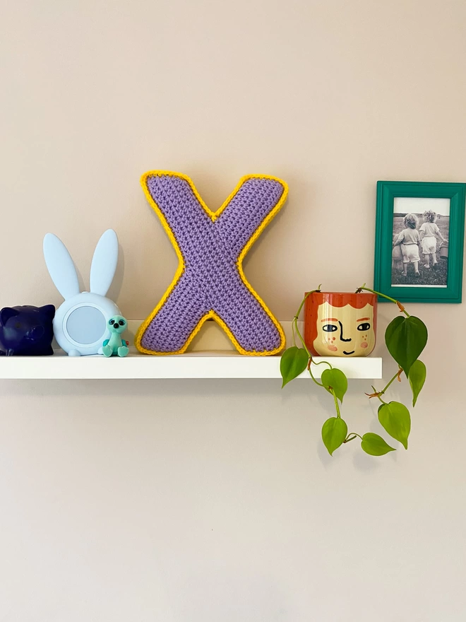 Crochet Cushion shaped like an X in Lilac and Sunshine Yellow, on a shelf