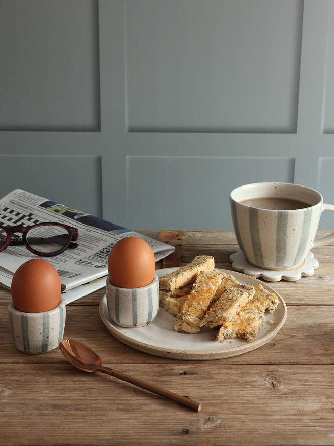 A breakfast scene featuring two handmade ceramic egg cups with a blue and white stripe design holding boiled eggs. One of the egg cups sits on a white handmade ceramic plate filled with buttered toast which sits in front of a handmade blue and white striped mug filled with coffee. A brass spoon and a newspaper with a pair of glasses on top also sit on the table. 