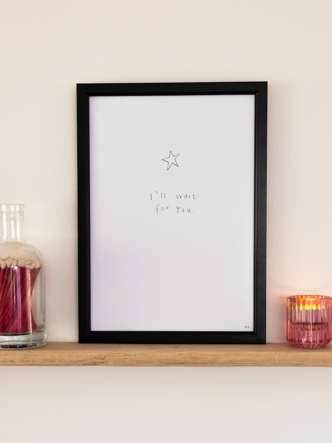 A simple line drawing star print in a black frame on a shelf next to a bottle of matches and a lit candle.