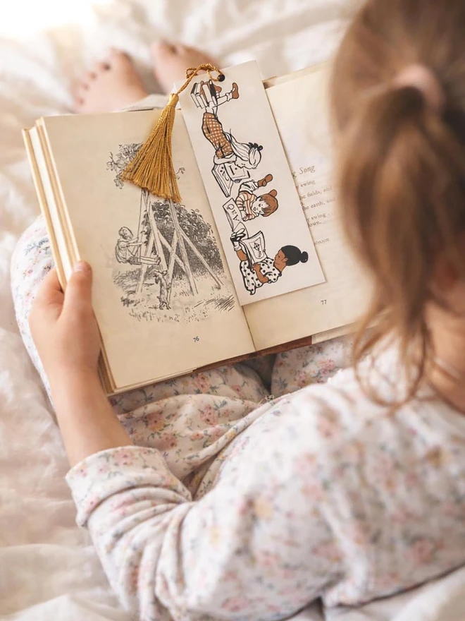 A young girl reading a story book with a beautifully illustrated book mark. The book mark is a traditional card bookmark with a golden brown swooshy tassel featuring an illustration of children reading