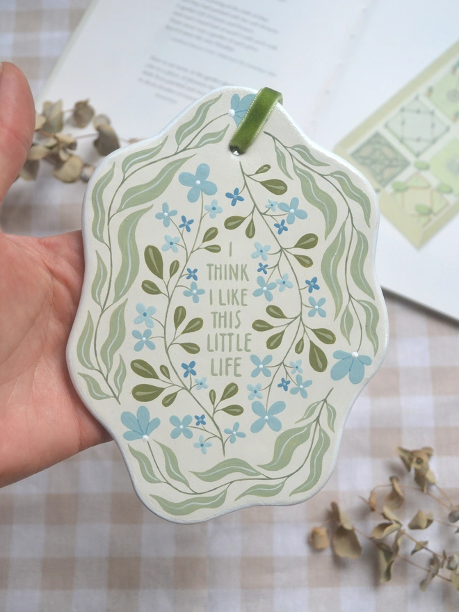 hanging clay sign with blue and green floral design reading 'i think i like this little life' in front of gingham background