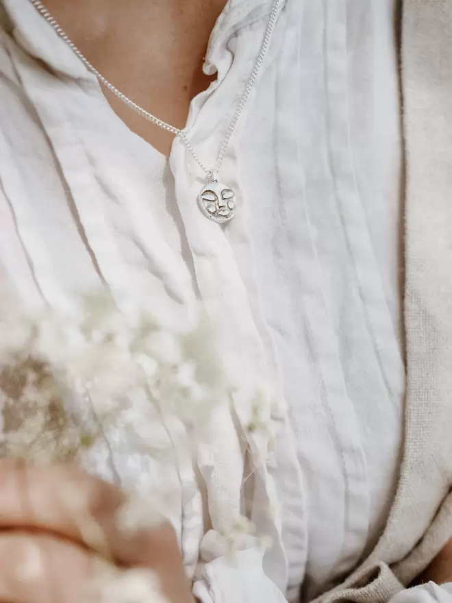 Image of woman’s chest wearing a white blouse and a hand carved sterling silver moon face pendant. Woman also holding a bunch of babies breath flowers which are at the forefront of the image but out of focus 