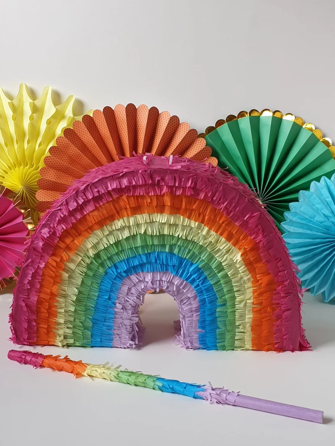 rainbow pinata with matching whacking stick in front of a rinbow of paper fans