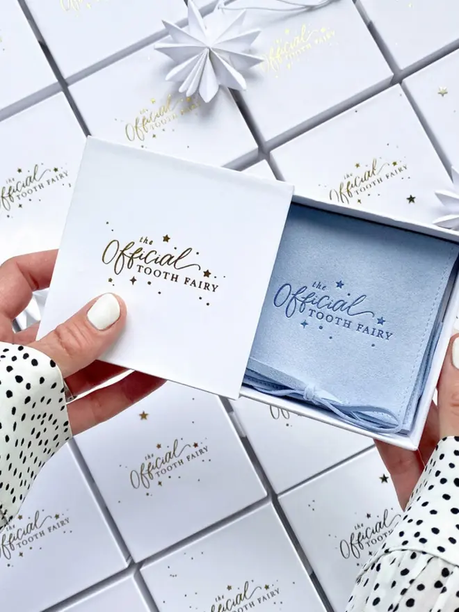 wo hands open a white gift box holding a pale blue suede pouch with the "Official Tooth Fairy" logo in blue. Rows of matching boxes fill the background.