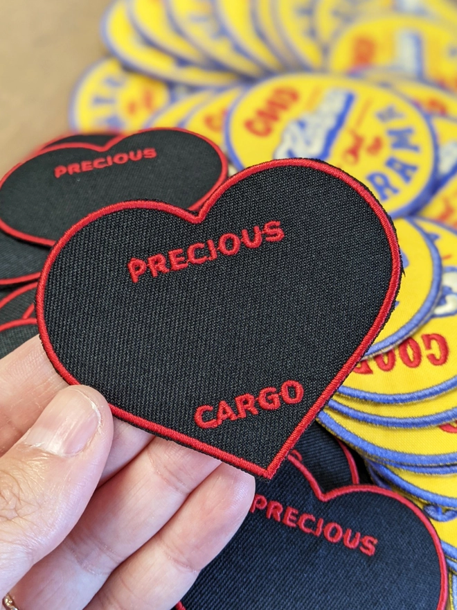 Precious Cargo Woven embroidered patch template, left blank of customisation. Black heart shaped patch with Red border, held up by hand in front of a bunch of patches on a table, reading 'Precious Cargo' in Red. 