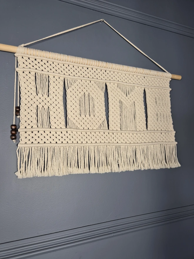 Macrame wall hanger featuring the word 'HOME' knotted in macrame square knots, perfect for enhancing your wall, space and ambience