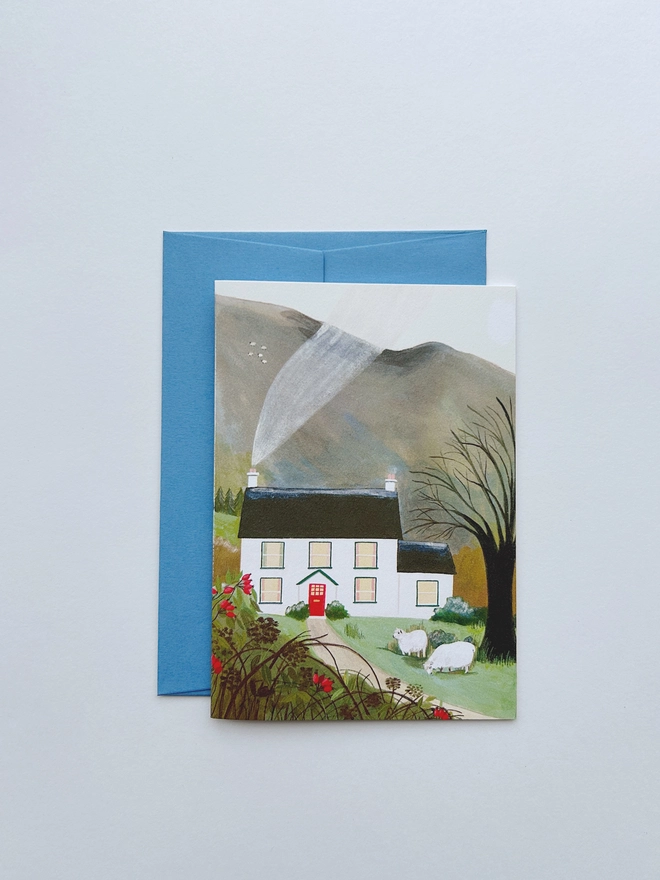 An illustrated greeting card. A painting of a white cottage nestled within trees against a backdrop of a mountain. The cottage had a red door and smoke curling from the chimney. At the front of the house their are two sheep happily grazing.