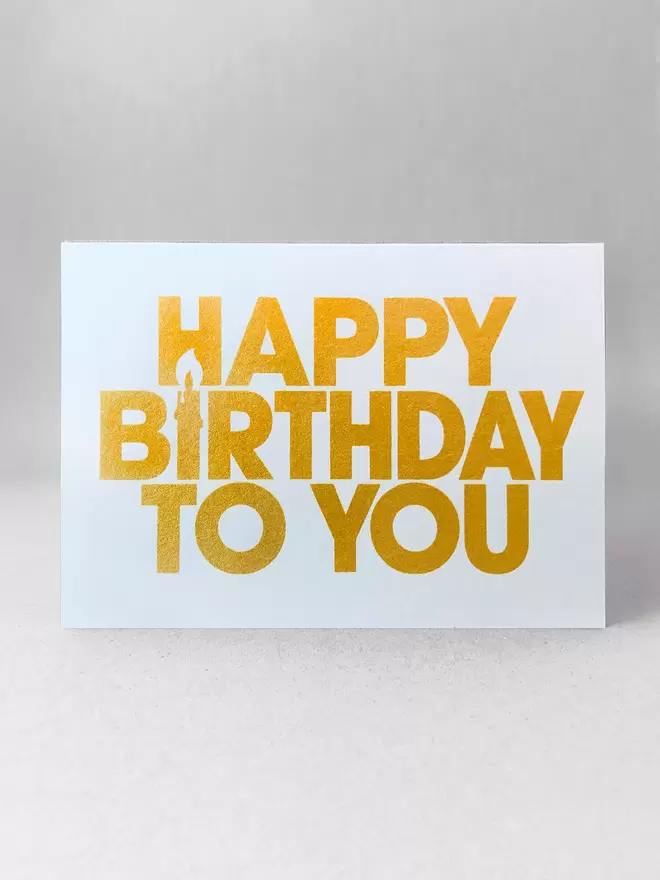 Happy Birthday to You landscape greetings card. The I of Birthday is a candle using negative space. A front on view - Printed in capital letters, simple font printed in a gold ink on a white card made by Salty's Studio.