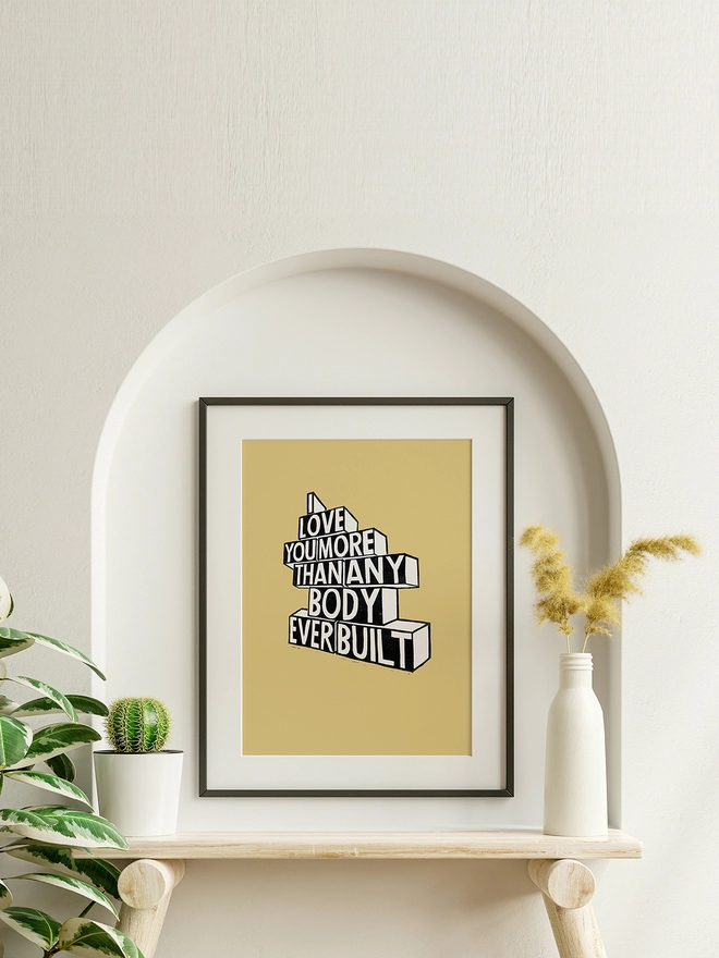 A framed yellow Woodism print hung inside an alcove on a wall. The typography reads: I Love You More Than Anybody Ever Built.