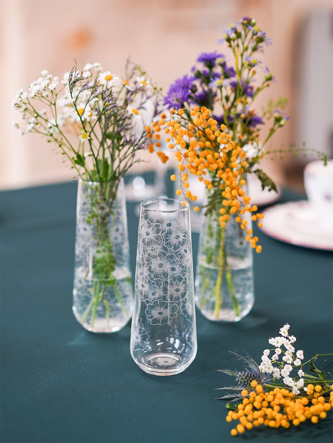 Three clear Floral Twist crystal bud vases holding colourful wildflower arrangements placed on a dark green tablecloth.