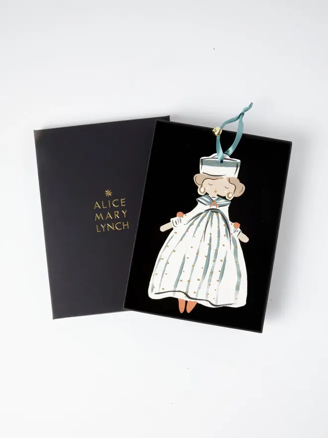 Sailor Queen Tree Dancer. the sailor queen tree dancer hanging ornament sits inside its black presentation box, with the lid open to reveal "alice mary lynch" embossed in gold lettering. the figure has curled hair, gold hoop earrings and a striped sailor hat, wearing a teal and cream striped gown dotted with gold. the branded box gives the piece a polished, gift-ready presentation.
