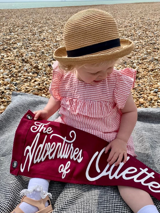 A little girl sat on the beach holding a The adventures of pennant flag