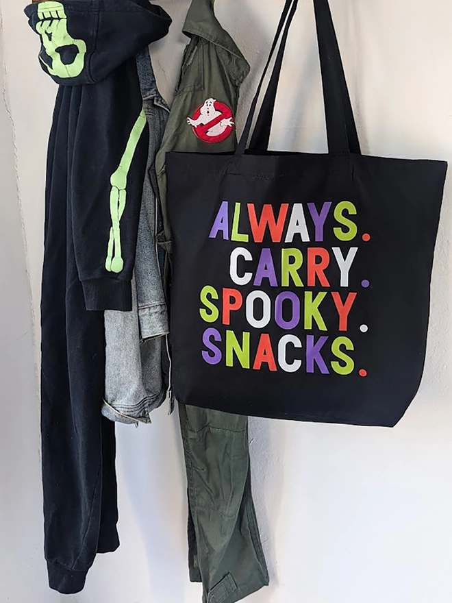 A black tote bag with multi coloured Always Carry Spooky Snacks slogan.