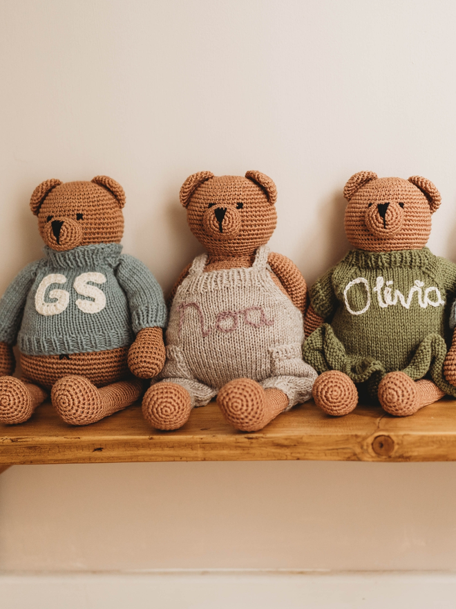 three hand crochet brown teddy bears on wooden shelf wearing hand knit personalised clothing