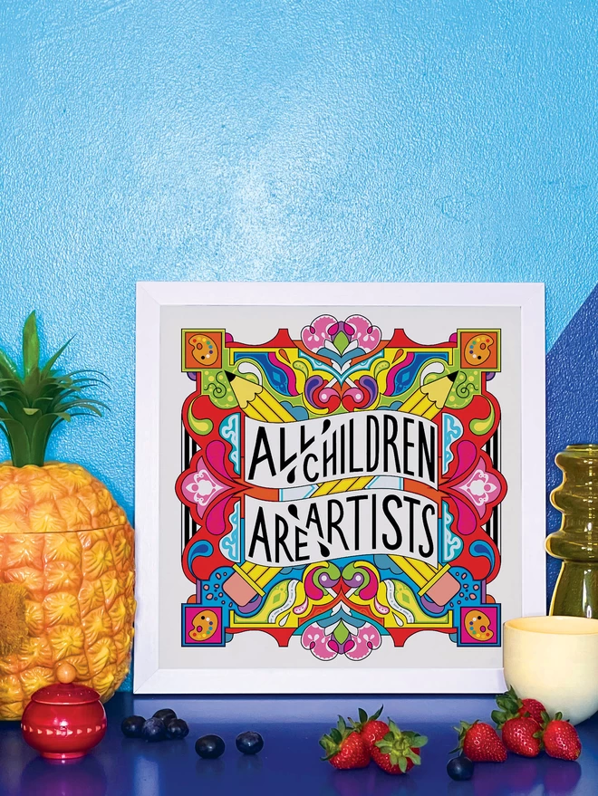 A white square frame surrounds the print, which has the words All Children are Artist at the centre of this multi-coloured vibrant illustration which includes a cross of pencils, and artists palettes the corners. It is propped against a wall painted light and dark blue, on a shiny blue cabinet. Next to the frame is a plastic pineapple ice bucket, a small wooden red pot a handful of blueberries, a yellow glass vase, a small yellow ceramic pot and 4 strawberries.