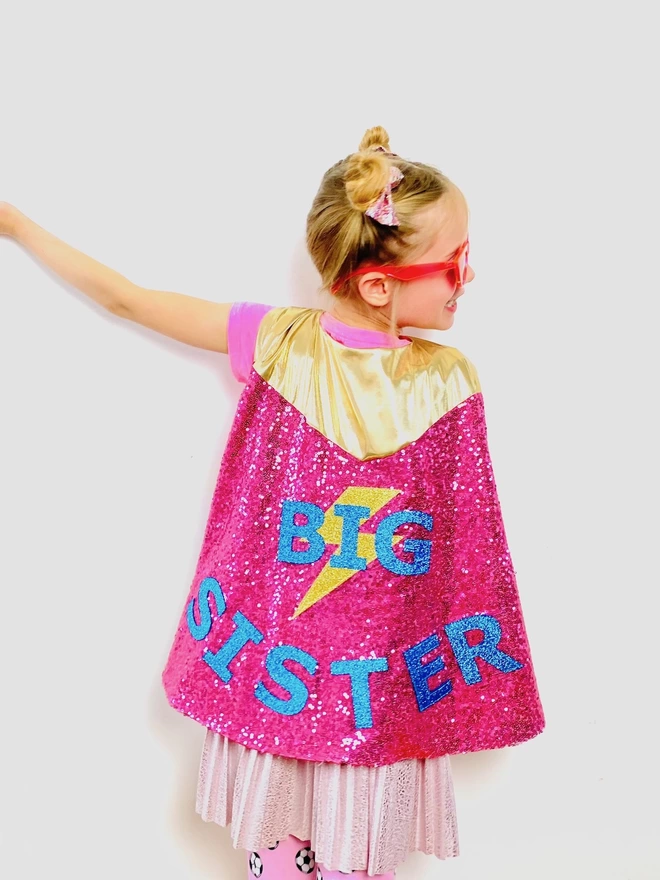 The back of a young girl wearing the big sister cape.  It has pink sequins and a gold neck fastening. The text reads 'BIG SISTER' in blue, with a gold lightening bolt layered underneath the word 'BIG'