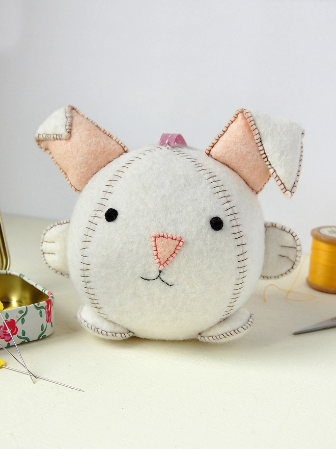 A handmade white felt rabbit toy with brown stitching stands on a white desk beside various craft kit components.