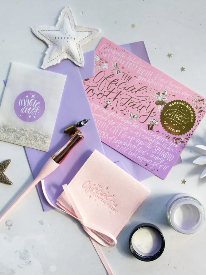 The pink-lilac notecard lies on a lilac envelope beside a blush pink pouch and a lilac Magic Dust packet. A rose gold dip pen and a "Believe" paper star complete the scene.