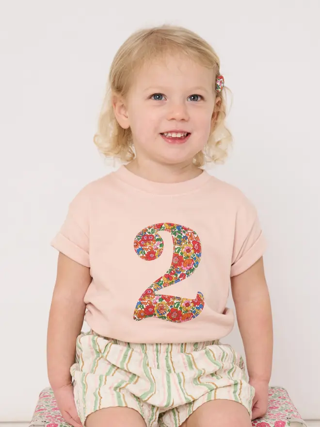 A smiling 2 year old girl wearing a number pink t-shirt on her birthday featuring a floral liberty number 2 on the front.
