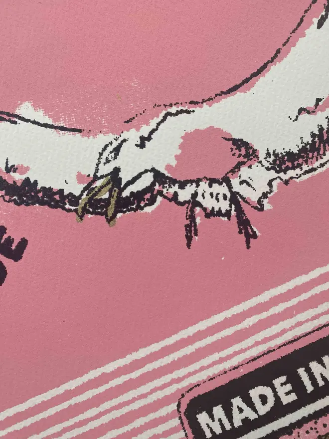 
A close-up photograph of a limited edition A2 screenprint. The print depicts a retro-style matchbox illustration in pink, black, and gold. A large, realistic seagull with a mischievous expression and an open beak is flying across the center. The words "THIEVING SEAGULL" are at the top, and "TRADE MARK" is on either side of the seagull. A banner at the bottom says "MADE IN FOLKESTONE." The background is a solid pink with a slight gold texture. The matches on the side are green and heart-shaped. Below the main image, the print is signed and numbered 3/13. The entire image has a grainy texture, like a vintage screen print.