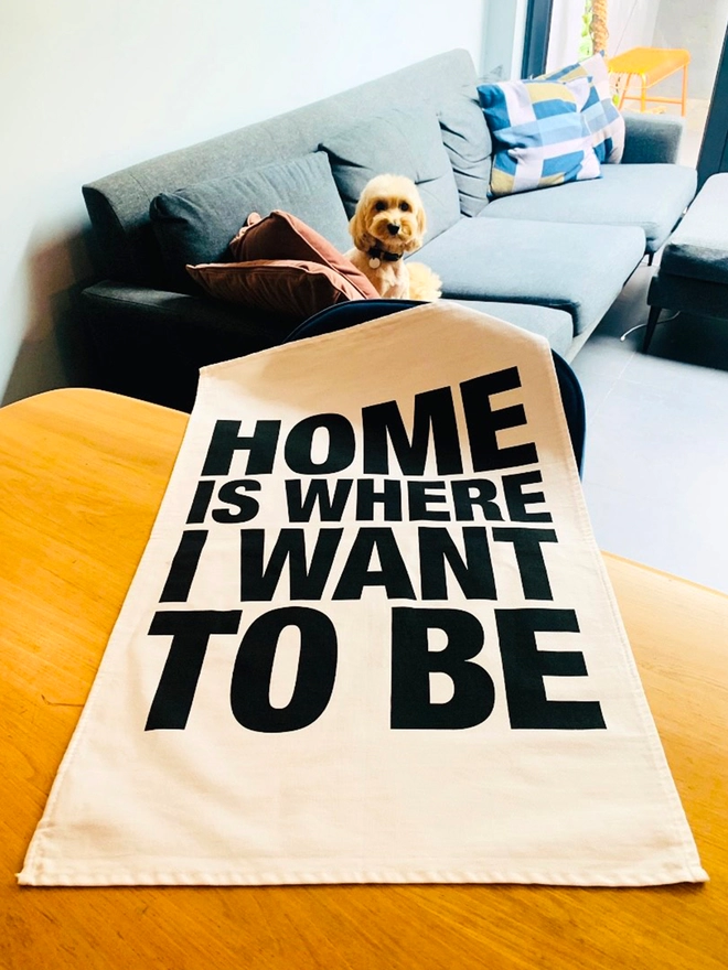 London Drying Home Is Where I Want to Be black screen printed text on white tea towel laying on timber table with dog sat on sofa in background