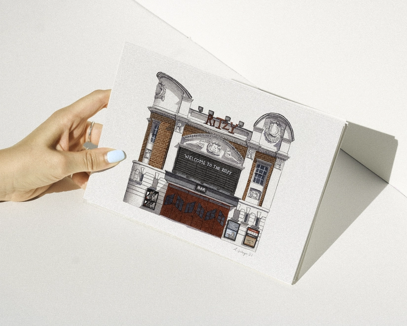 A hand with blue nail polish holding a print featuring an illustration of the Ritzy Cinema against a white background.