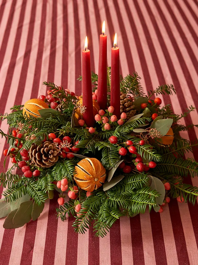 green wreath with dried oranges, red berries, and leaves on a table with red striped table cloth. three lit, red candles in the middle