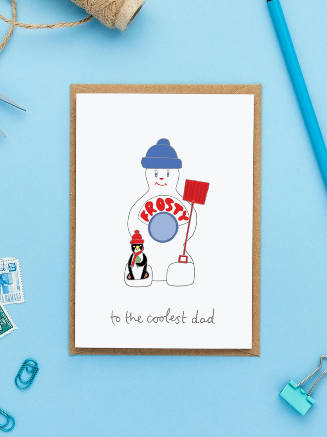Greeting card featuring a hand-drawn Frosty ice lolly character with a red shovel and a penguin, styled on a blue background with twine, pencil and kraft envelope.