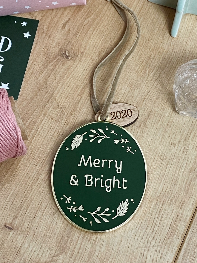 A deep green and gold enamel Christmas decoration, with the words Merry & Bright surrounded by golden flowers, lays on a wooden desk.