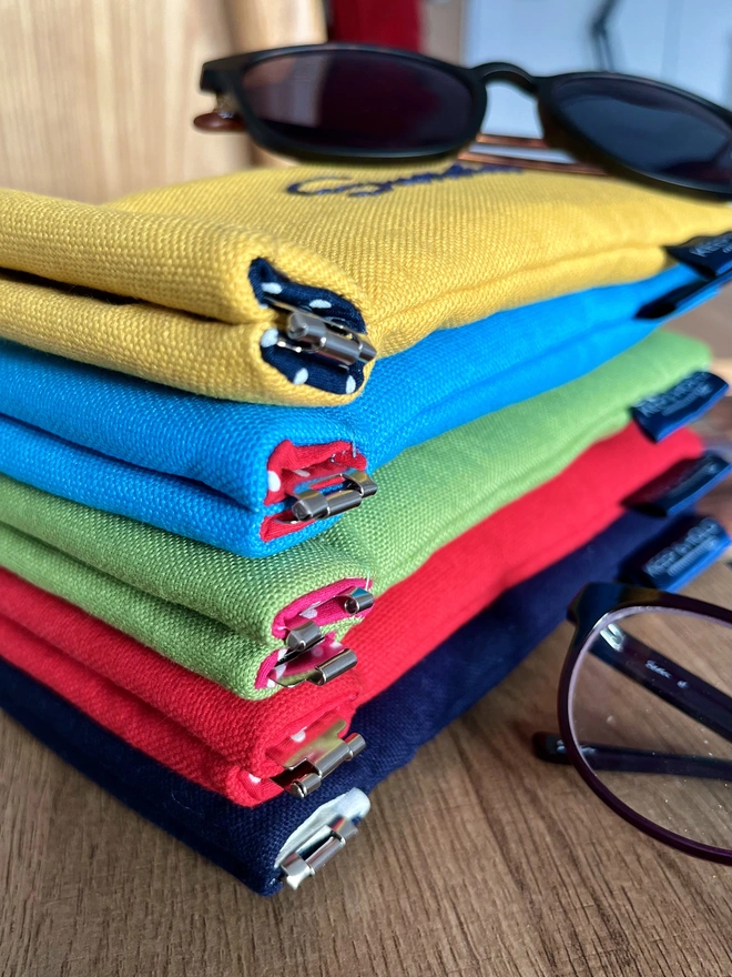 Stack of colourful cotton glasses cases. Sunglasses & a pair of spectacles laying on top and beside.