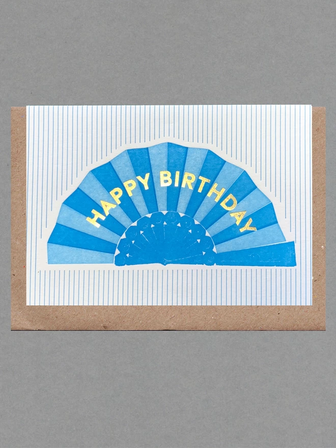 White card with blue fan with gold text reading 'Happy Birthday' on blue striped background with brown envelope behind