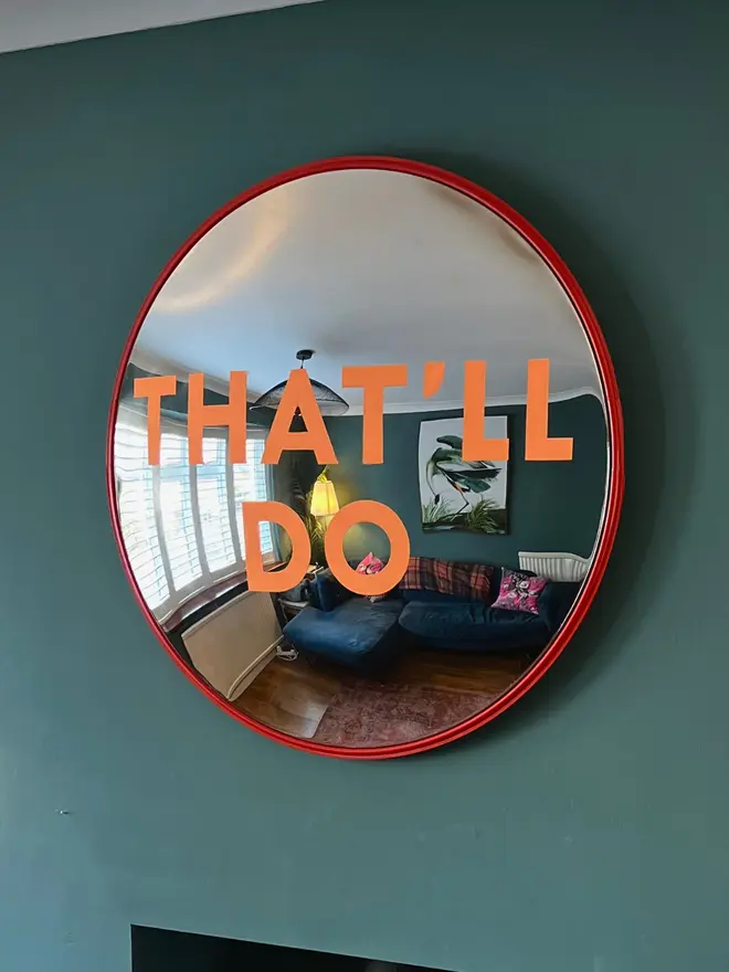 Round convex mirror with 'That'll Do' hand painted in orange in a red round frame, on a teal wall and log burner, with a lamp, logs and white ceiling