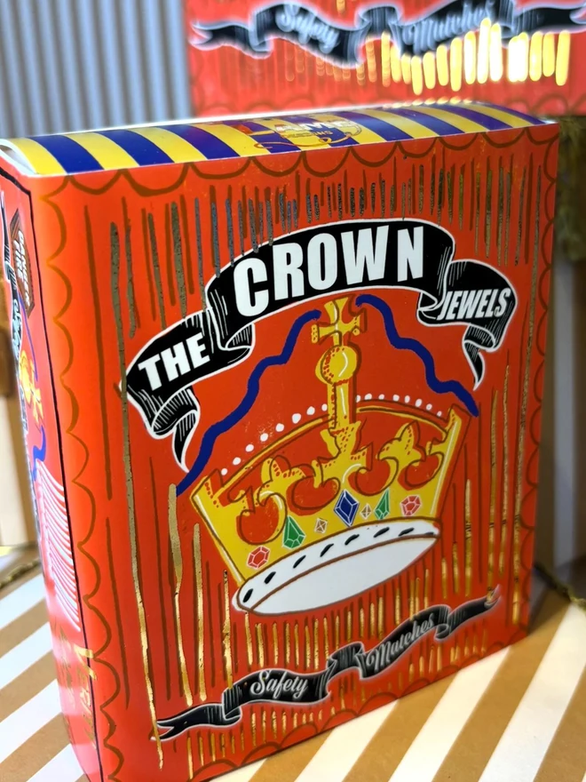 The Crown Jewels Matchbox, a red matchbox with a crown design sitting on a decorated surface accompanied by other accessories. 