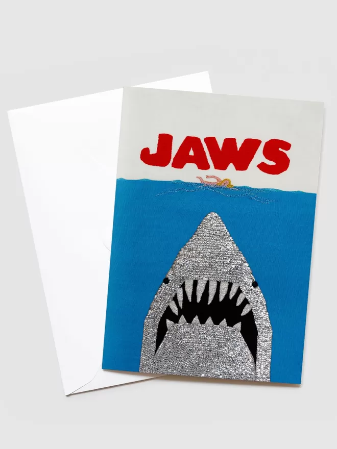 Kate Jenkins Stitch Attack Blank Greetings Card seen with envelope. On the front of the card is a hand stitched silver shark with pointed teeth looking up towards a swimmer in the sea. Jaws is stitched above in bold red letters. 