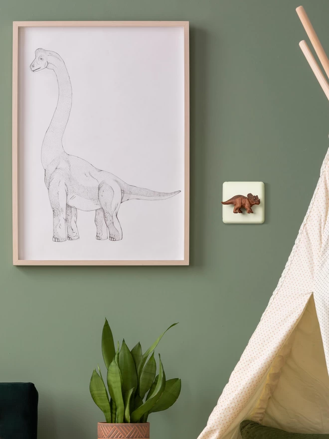 A dinosaur light switch on a sage green painted wall next to a line drawing of a brachiosaurus in a wooden frame. A brown triceratops dinosaur is used as the rotary dimmer switch knob on the light switch. The light switch plate is cream and made of metal, epoxy coated steel. The triceratops is made of plastic. The brand is Candy Queen Designs. There is a cream teepee to the right and a plant in a terracotta vase.