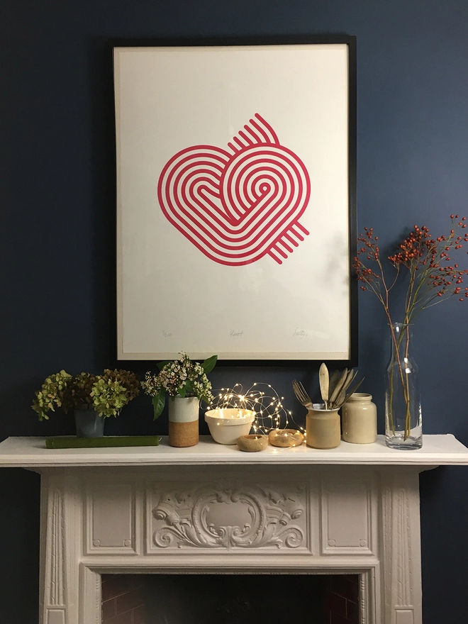 Stripy graphic magenta heart screenprint design, in a black frame hung on a dark blue wall above a white Victorian fireplace decorated with bowls, flowers and fairy lights.