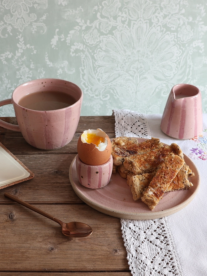 Two pink-striped ceramic egg cups holding boiled eggs sit on a wooden table alongside a matching plate of buttered toast, a mug and a milk jug. 